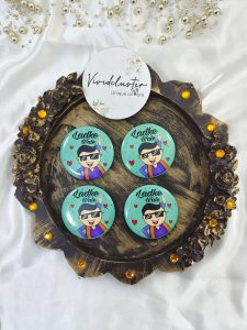 displaying a mint-green brooch with a cartoon figure in sunglasses and traditional outfit, text "Ladke Wale." Several matching brooches are scattered behind Great for baraat day, nikah ceremonies, or as unique wedding favors.