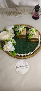 A beautifully crafted ring ceremony platter featuring a green and gold fabric base with white pearls and artificial flowers. It includes two gold boxes, one with an open red velvet interior, and is decorated with gold beads