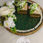 A beautifully crafted ring ceremony platter featuring a green and gold fabric base with white pearls and artificial flowers. It includes two gold boxes, one with an open red velvet interior, and is decorated with gold beads