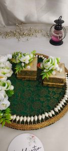 An ornate ring ceremony platter designed with a green and gold base, white and green flowers, and a pearl border. It has two gold boxes, one open with a red velvet lining, ng ceremony platter combines elegance and tradition