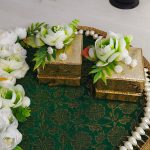 An ornate ring ceremony platter designed with a green and gold base, white and green flowers, and a pearl border. It has two gold boxes, one open with a red velvet lining, ng ceremony platter combines elegance and tradition