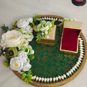 A decorative ring ceremony platter adorned with white and green artificial flowers, featuring a green fabric base with a gold pattern, bordered by white pearls. The platter includes two small gold boxes, one with an open red velvet interior