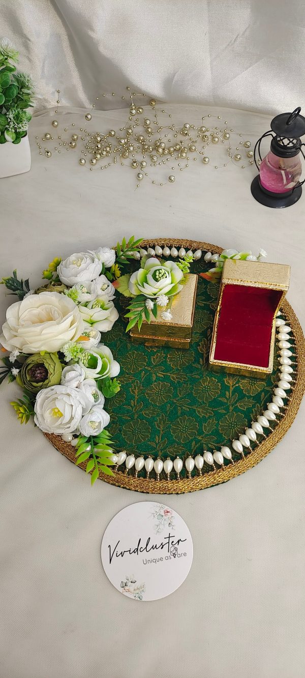 A decorative ring ceremony platter adorned with white and green artificial flowers, featuring a green fabric base with a gold pattern, bordered by white pearls. The platter includes two small gold boxes, one with an open red velvet interior