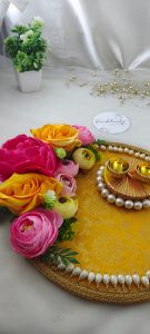 decorative wedding Roli Kumkum Pooja platter ideal for wedding or nikah ceremonies. It features a bright yellow fabric base with a gold floral pattern, bordered by white pearls. Adorned with pink, yellow, and peach artificial flowers, the platter includes two small golden cups on a circular pearl arrangement.