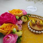 decorative wedding Roli Kumkum Pooja platter ideal for wedding or nikah ceremonies. It features a bright yellow fabric base with a gold floral pattern, bordered by white pearls. Adorned with pink, yellow, and peach artificial flowers, the platter includes two small golden cups on a circular pearl arrangement.