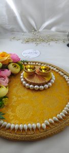 A vibrant wedding Roli Kumkum Pooja platter with a yellow fabric base, decorated with pink, yellow, and peach artificial flowers, and bordered by white pearls. It includes two small golden cups on a circular arrangement of pearls.