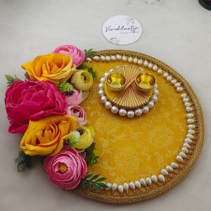 Roli Kumkum Pooja Platter wedding and nikah ceremonies. The platter features a bright yellow fabric base with intricate floral patterns, bordered by white pearls. Adorned with vibrant artificial flowers in shades of pink, yellow, and peach, the platter includes two small golden cups placed on a circular arrangement of pearls, perfect for holding roli and kumkum or rings