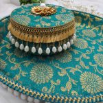 a round decorative laddu box with a matching bangle ceremony multipurpose tray, both adorned in a turquoise fabric with intricate gold floral patterns. The box is topped with a golden flower embellishment and a single pearl at its center. The edges are trimmed with golden tassels and hanging pearl beads