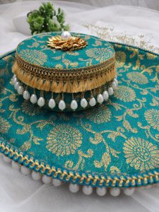 a round decorative laddu box with a matching bangle ceremony multipurpose tray, both adorned in a turquoise fabric with intricate gold floral patterns. The box is topped with a golden flower embellishment and a single pearl at its center. The edges are trimmed with golden tassels and hanging pearl beads