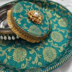 Decorative laddu box with a coordinating shagun decorative tray, wrapped in turquoise fabric with gold floral designs. The box has a golden flower with a pearl on top and is bordered by golden tassels and pearl beads. The tray has a matching blue and gold braided trim decorative wedding platter