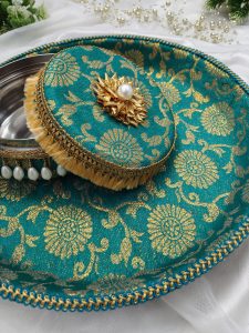 Decorative laddu box with a coordinating shagun decorative tray, wrapped in turquoise fabric with gold floral designs. The box has a golden flower with a pearl on top and is bordered by golden tassels and pearl beads. The tray has a matching blue and gold braided trim decorative wedding platter