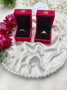 Ring ceremony platter with red velvet ring box, placed on a white satin cloth adorned with pearls. The setup includes red roses, greenery, and gold bead decorations, ideal for wedding jewelry presentations ring ceremony platter