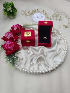 Beautiful wedding ring ceremony setup with a gold ring in a red velvet box on a pearl-trimmed satin cloth, enhanced by red roses and greenery attracting brides-to-be with its elegant presentation