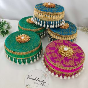Five beautifully decorated round Wedding giveaway laddu boxs in vibrant colors (green, blue, and pink) with intricate gold patterns, adorned with pearl tassels and golden floral designs boxes are ideal for storing and presenting shagun laddus