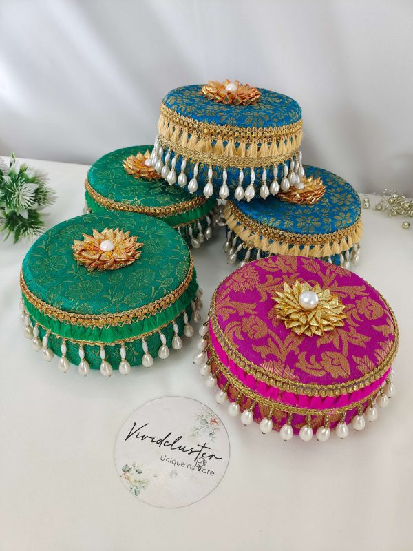 Five beautifully decorated round Wedding giveaway laddu boxs in vibrant colors (green, blue, and pink) with intricate gold patterns, adorned with pearl tassels and golden floral designs boxes are ideal for storing and presenting shagun laddus
