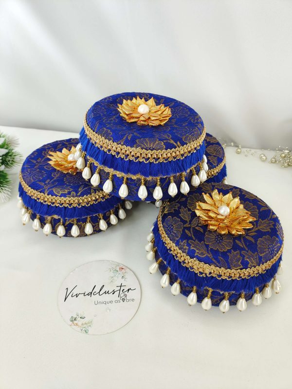 Four blue round laddu boxes featuring golden floral patterns and golden floral motifs with pearls. The boxes are elegantly embellished with golden lace and hanging white pearl beads, perfect for wedding gifts and ceremonies