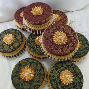 A collection of ornate round laddu boxes in maroon and green with intricate gold floral patterns, adorned with golden floral embellishments and pearl trimmings for presenting to guests and baratis as memorable wedding