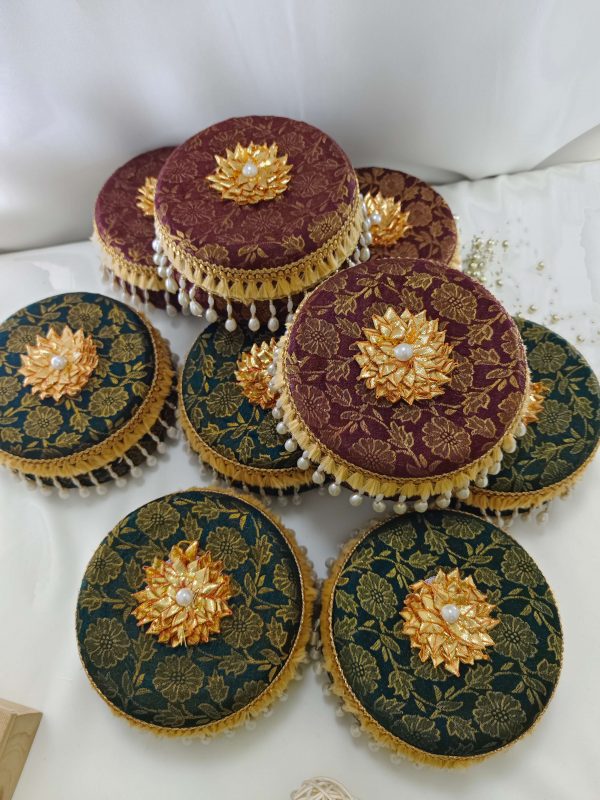 A collection of ornate round laddu boxes in maroon and green with intricate gold floral patterns, adorned with golden floral embellishments and pearl trimmings for presenting to guests and baratis as memorable wedding