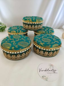 Vibrant teal, green, yellow, and white round shagun boxes with intricate gold detailing and pearl accents, perfect for wedding favors