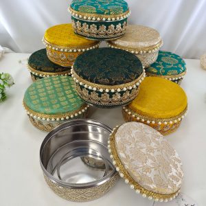 A collection of beautifully decorated round wedding laddu shagun boxes in various colors including teal, green, yellow, and white, adorned with intricate gold embroidery and pearl accents presenting wedding favors, barati gifts, and bridesmaids' shagun return gift