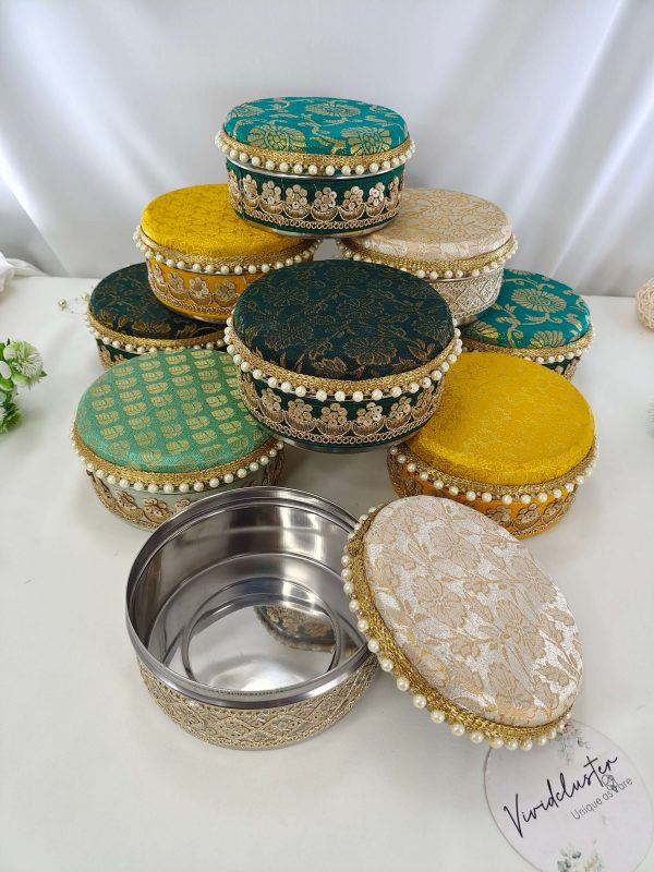 A collection of beautifully decorated round wedding laddu shagun boxes in various colors including teal, green, yellow, and white, adorned with intricate gold embroidery and pearl accents presenting wedding favors, barati gifts, and bridesmaids' shagun return gift