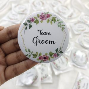 a round Team Groom Badges with "Team Groom" written in elegant black script. The brooch is adorned with pink and green floral designs at the top and bottom This wedding accessory is perfect for groomsmen at a nikah ceremony, adding a touch of elegance to their wedding attir