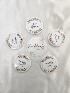 a round brooch with "Team Groom" written in elegant black script. The brooch is adorned with pink and green floral designs at the top and bottom This wedding accessory is perfect for groomsmen at a nikah ceremony, adding a touch of elegance to their wedding attir