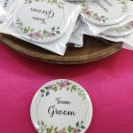 "Team Groom" brooch held between fingers. The brooch features pink and green floral decorations on a white base for groomsmen at a wedding or nikah ceremony