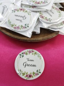 "Team Groom" brooch held between fingers. The brooch features pink and green floral decorations on a white base for groomsmen at a wedding or nikah ceremony