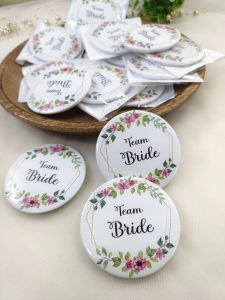 pink and white team bride brooch featuring the text team bride with a floral print accessory for bride's side members during wedding ceremonies, baraat day, or nikah events