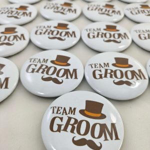 A collection of white circular brooches featuring the text "TEAM GROOM" in brown, adorned with a brown top hat and mustache These brooches serve as great wedding favors, baarati brooches, and wedding attire accessories.
