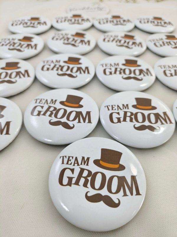 A collection of white circular brooches featuring the text "TEAM GROOM" in brown, adorned with a brown top hat and mustache These brooches serve as great wedding favors, baarati brooches, and wedding attire accessories.